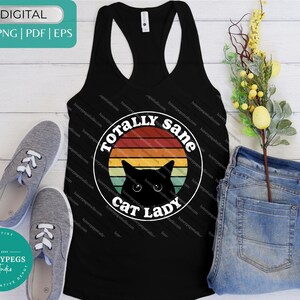Könnte beinhalten: Schwarzes Tanktop mit einem Aufdruck "TOTALLY sane cat lady" in einem weißen Kreis mit Retro-Sonnenuntergangsdesign und einer schwarzen Katzen-Silhouette. Graue Turnschuhe und blaue Jeans liegen neben dem Tanktop.
