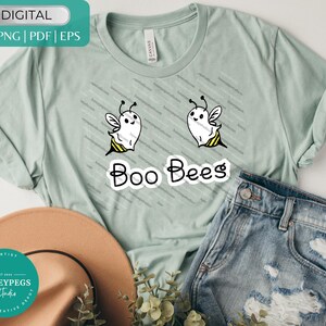 May include: A light green t-shirt with a "Boo Bees" graphic. The graphic features two cartoon bees with ghost-like bodies, white with black antennae and yellow and black stripes. The shirt is paired with blue denim shorts and a brown hat.