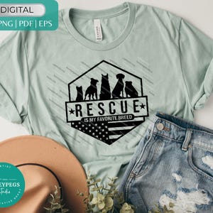 Op de afbeelding: Lichtgroen t-shirt met de tekst "RESCUE IS MY FAVORITE BREED" en een afbeelding van hondensilhouetten. Het ontwerp bevat een Amerikaans vlagmotief. Het shirt wordt gecombineerd met een blauwe jeans en een bruine hoed.