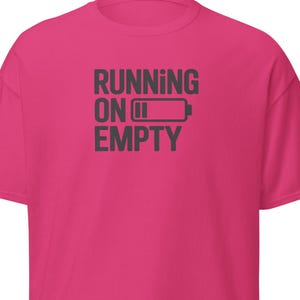 May include: A bright pink t-shirt with the phrase "RUNNING ON EMPTY" printed in dark gray. The design includes a battery icon indicating low power. The shirt is a short-sleeved, casual style.