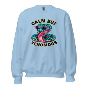 May include: Light blue sweatshirt featuring a cartoon snake wearing sunglasses. The snake is teal with pink accents and the text "CALM BUT VENOMOUS" in black and yellow. A crew neck sweatshirt.