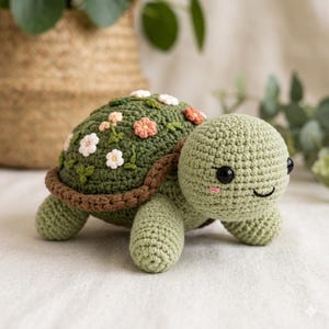 May include: A crocheted turtle in shades of green, with a brown shell border. The turtle's shell is adorned with white, pink, and orange crocheted flowers and green leaves. The turtle has black eyes and a small pink smile. The turtle is a handmade item.