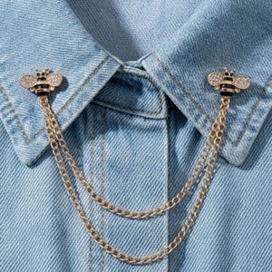 May include: A gold-toned metal collar pin set featuring two bee-shaped brooches with black enamel stripes and clear crystal wings. The two bee pins are connected by two delicate gold-toned metal chains that drape across the denim fabric.