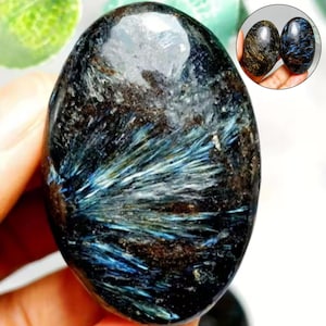 May include: An oval-shaped polished gemstone featuring a dark black base with radiating, needle-like inclusions in shades of metallic blue and silver. The stone has a smooth, glossy surface with a prominent white crystalline patch at the top.