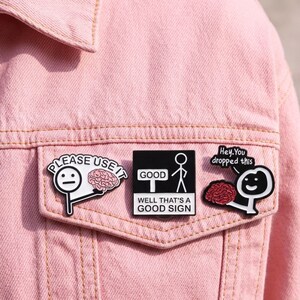 May include: Three enamel pins attached to a light pink denim jacket. The left pin is a white stick figure holding a pink brain with the text PLEASE USE IT. The centre square pin shows a stick figure and a sign reading GOOD WELL THAT'S A GOOD SIGN. The right pin is a black stick figure holding a red brain with the text Hey. You dropped this.