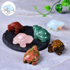 Puede incluir: Una colección de figuritas de tortugas de piedras preciosas en varios colores, incluyendo rojo, verde, rosa y un tono opalescente translúcido. Las tortugas se exhiben sobre una superficie oscura y texturizada, mostrando sus detalles tallados y patrones únicos. Las medidas varían de 2,5 a 4,3 cm.