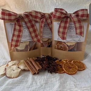 May include: Three brown paper pouches labeled Harvest Blend Stovetop Potpourri, each topped with a red and white gingham ribbon. The pouches contain dried orange slices, cinnamon sticks, and star anise, which are also displayed in front.