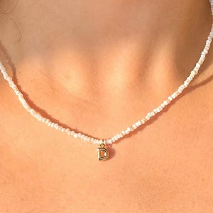Peut inclure: Collier de perles blanches avec un pendentif doré en forme de la lettre "D". Le collier est composé de petites perles blanches rondes. Le pendentif est centré et suspendu au collier.