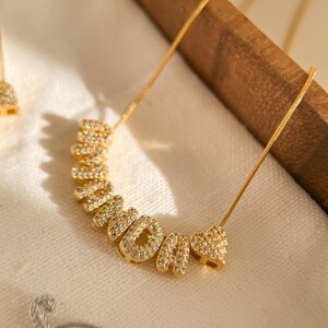 Può includere: Una collana color oro con la scritta "MOMDA" in lettere scintillanti e tempestata di gemme. Le lettere sono disposte in una linea curva. La collana è su una catena delicata. Lo sfondo è un morbido tessuto bianco.
