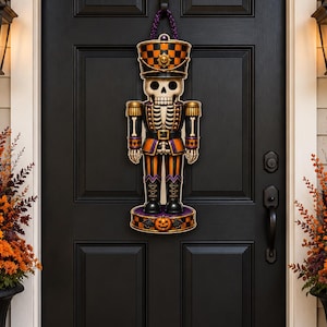 May include: A hanging door decoration shaped like a skeleton nutcracker. The figure features a skull head, a checkered orange and black hat with a skull emblem, and a uniform in orange, black, and purple. A small pumpkin sits on the base.