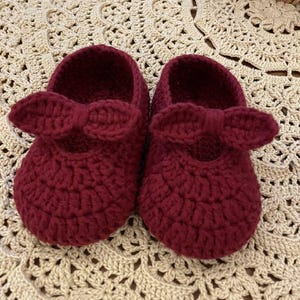May include: A pair of burgundy crochet baby shoes with bow accents. The shoes are handmade and sit on a cream-colored, patterned surface. The text "HANEULWOOLSHOP" is at the bottom of the image.