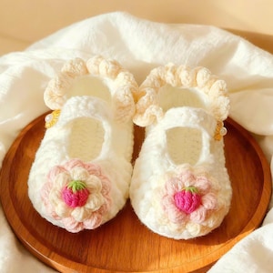 Puede incluir: Un par de zapatos de bebé de ganchillo blanco hechos a mano con un diseño floral. Los zapatos presentan una flor rosa y verde en la puntera y un ribete con volantes. El texto "HANEULWOOLSHOP" está en la parte inferior.