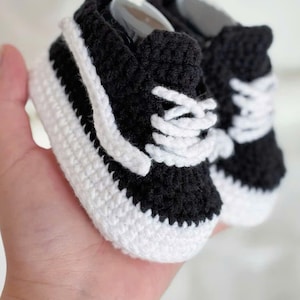 May include: Hand-crocheted baby sneakers in black and white. The tiny shoes feature a classic sneaker design with white laces and a white stripe on the side. The soles and the stripe are white, while the rest of the shoe is black.