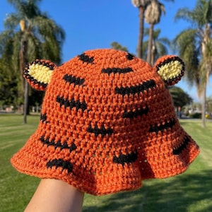 May include: Orange and black crochet bucket hat with tiger stripes and yellow ear accents. The hat features a wide brim and is handmade. The text "HANEULWOOLSHOP" is at the bottom of the image.