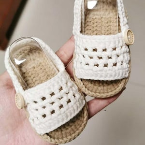 Może przedstawiać: Para ręcznie robionych sandałów dla niemowląt. Sandały są szydełkowane z białą górą i brązową podeszwą. Każdy sandał ma mały drewniany guzik i przezroczyste plastikowe podparcie łuku. Sandały są prezentowane na dłoni.