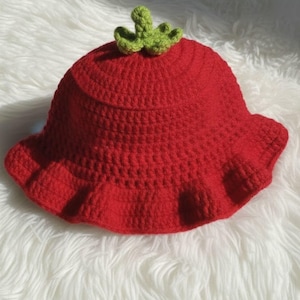 May include: A handmade red crochet hat with a ruffled brim and a green leaf detail on top. The hat is displayed on a white, fluffy surface. The text "HANEULWOOLSHOP" is at the bottom.