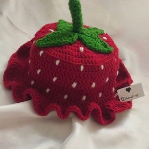 May include: A red crochet strawberry hat with white seed details, a green stem and leaves, and a ruffled edge. The handmade hat is designed to look like a strawberry. The shop name "HANEULWOOLSHOP" is at the bottom.