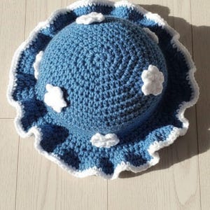 May include: A handmade blue crochet hat with a white scalloped edge and white cloud accents. The hat is round with a wide brim and is sitting on a light wood surface. The text "HANEULWOOLSHOP" is at the bottom.