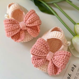May include: A pair of handmade, crocheted baby booties in cream and peach colors. Each bootie features a large, decorative bow with a velvet ribbon accent. The booties are adorned with a small pearl detail. The text "HANEULWOOLSHOP" is visible.