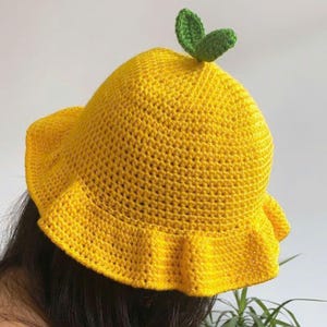 Pode incluir: Chapéu amarelo de crochê com uma borda franzida e uma decoração de folhas verdes. O chapéu é de um amarelo vibrante e parece ser feito de fio macio. O texto "HANEULWOOLSHOP" é visível na parte inferior.