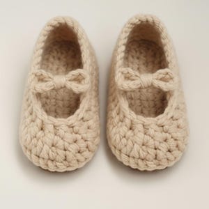 May include: A pair of cream-colored, crocheted baby shoes. Each shoe features a small bow on the front and a Mary Jane style strap. The shoes are handmade and appear soft and comfortable. The text "HANEULWOOLSHOP" is at the bottom.