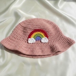 Op de afbeelding: Een roze gehaakte vissershoed met een regenboog applicatie. De regenboog heeft rode, oranje, gele, groene en blauwe bogen met witte wolken accenten. De hoed wordt gepresenteerd op een witte satijnen ondergrond. De tekst "HANEULWOOLSHOP" staat onderaan.