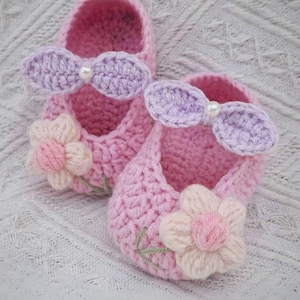 May include: A pair of handmade pink crochet baby shoes. Each shoe features a light purple bow with a pearl accent and a light pink and white flower. The shoes are displayed on a white textured surface. The text "HANEULWOOLSHOP" is at the bottom.