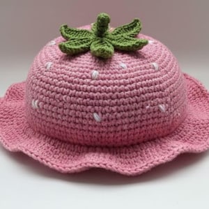 May include: A pink crocheted hat shaped like a strawberry, featuring a green stem and leaves on top. The hat has white stitches and a scalloped brim. The text "HANEULWOOLSHOP" is at the bottom.