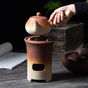 以下が含まれることがあります： 茶色のグラデーションとクリーム色のセラミックティーセット。セットにはティーポットと小さな開口部のあるベースが含まれています。ティーポットは手で持たれています。セットは木製の表面にあります。