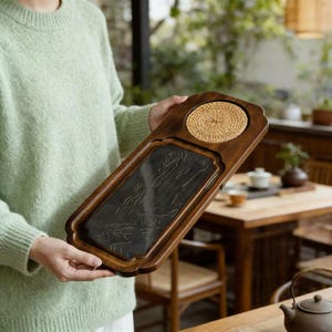 May include: A rectangular wooden tea tray with a dark, carved stone insert. The tray has a woven coaster and a decorative edge. The wood is a rich brown color, and the stone insert features an etched design. The tray is held by a person wearing a green sweater.