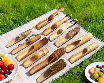 May include: A collection of handcrafted bamboo tea scoops, each with a unique design and varying shades of brown. The scoops are arranged on a light-colored cloth, with plates of fruit on either side, set against a backdrop of green grass.
