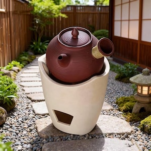 Pode incluir: Uma chaleira de cerâmica marrom com tampa e bico, repousando em uma base de cerâmica creme. A chaleira tem formato redondo com superfície texturizada. A base tem um design recortado. A cena é ambientada ao ar livre em um jardim japonês.