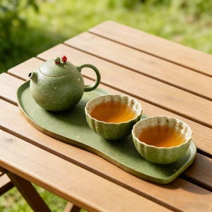 May include: A green ceramic tea set on a wooden table. The set includes a round teapot with a small handle and two cups, all filled with tea. The teapot has a small red and green decoration on top. The set sits on a green tray.