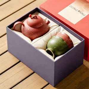 以下が含まれることがあります： ギフトボックスに入った陶器のティーセット。 赤茶色のティーポットと、お揃いのカップがセットになっており、どちらもまだら模様です。 カップは底が緑色で、上部が赤茶色です。 箱は紫で、赤い蓋と「DONG YANG」と書かれたラベルが付いています。