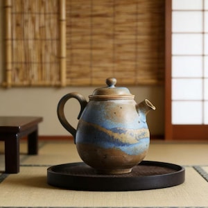 May include: A ceramic teapot with a rounded body, a curved handle, and a spout. The teapot has a speckled brown and blue glaze, and sits on a dark wooden tray. The background shows a traditional Japanese interior with bamboo blinds and a tatami mat.