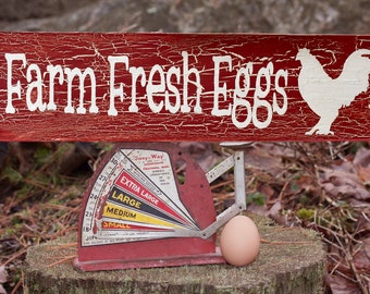 May include: A red wooden sign with white lettering that reads "Farm Fresh Eggs" with a white silhouette of a chicken. A red egg scale with a brown egg on the scale.