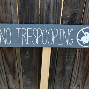 Può includere: Un cartello di legno grigio con testo bianco che recita "NO TRESPOOPING" e una silhouette bianca di un cane con un cerchio rosso e una linea diagonale che lo attraversa. Il cartello è attaccato a un palo di legno.