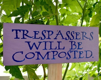 Puede incluir: Un cartel morado con letras azules que dice "Los intrusos serán compostados". El cartel está sujeto a una estaca de madera y está rodeado de plantas verdes frondosas.