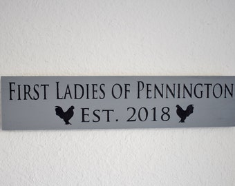 Puede incluir: Un letrero de madera gris con texto negro que dice "First Ladies of Pennington Est. 2018". El letrero tiene una silueta negra de un pollo a cada lado del texto.