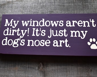 May include: A rectangular, purple wooden sign with white text that reads, "My windows aren't dirty! It's just my dog's nose art." A white paw print is in the lower right corner. The sign is on a gray wooden surface.