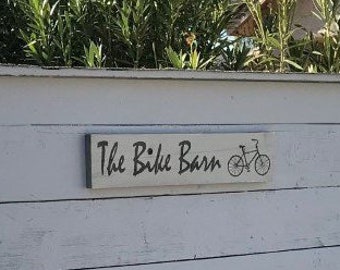 May include: A white wooden shed with a sign that reads "The Bike Barn" with a bicycle icon. The shed has horizontal wood planks.