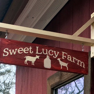 Puede incluir: Un letrero de madera rojo con letras blancas que dice "Sweet Lucy Farm". El letrero est&aacute; colgado de un soporte de madera sujeto a un edificio rojo. El letrero presenta una silueta de una cabra y una jarra de leche.