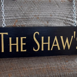 May include: Black wooden sign with gold lettering that reads "The Shaw's" hanging from a silver chain.