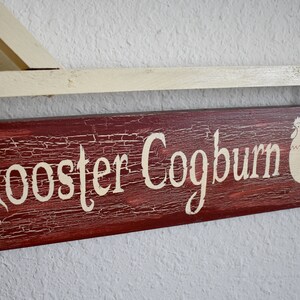 Puede incluir: Un letrero de madera r&uacute;stico con el texto "Rooster Coogburn" pintado en blanco sobre un fondo rojo. Una silueta de gallo blanca est&aacute; pintada en el lado derecho del letrero.