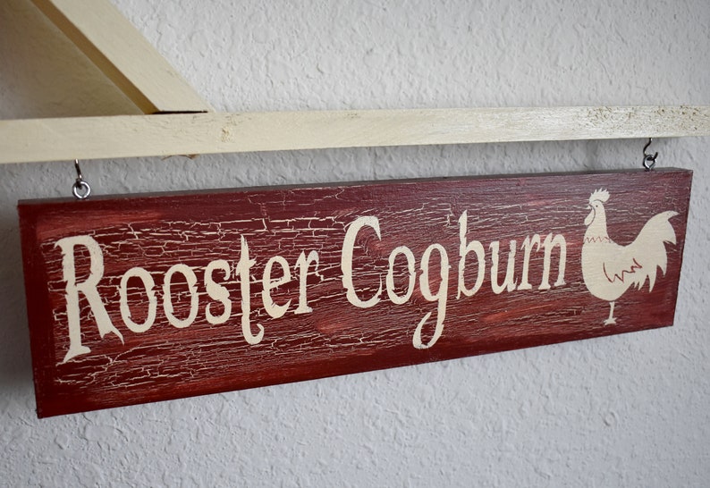 Puede incluir: Un letrero de madera r&uacute;stico con el texto "Rooster Coogburn" pintado en blanco sobre un fondo rojo. Una silueta de gallo blanca est&aacute; pintada en el lado derecho del letrero.
