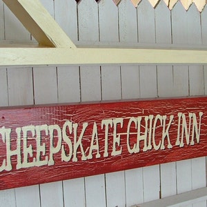 Puede incluir: Un letrero de madera rojo con letras blancas que dice "Cheapskate Chick Inn" y una silueta blanca de un pollo en el lado derecho. El letrero est&aacute; colgado de una valla blanca de madera.
