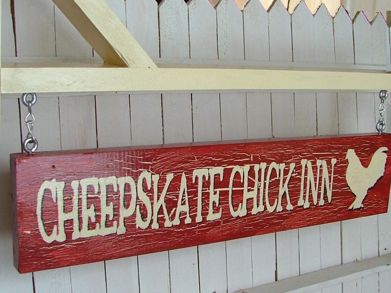 Puede incluir: Un letrero de madera rojo con letras blancas que dice "Cheapskate Chick Inn" y una silueta blanca de un pollo en el lado derecho. El letrero est&aacute; colgado de una valla blanca de madera.