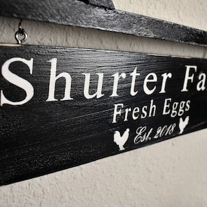 Puede incluir: Un letrero de madera negro con letras blancas que dice "Shurter Farm Fresh Eggs Est. 2018" con una silueta blanca de un pollo a cada lado del texto.