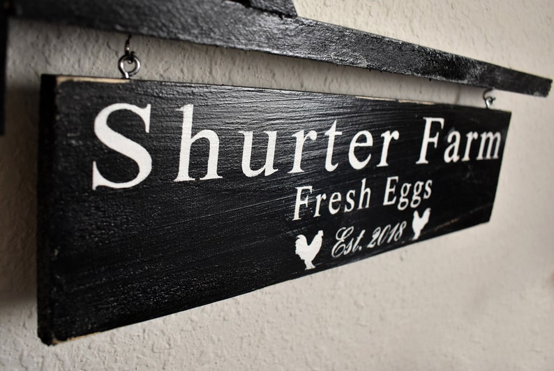Puede incluir: Un letrero de madera negro con letras blancas que dice "Shurter Farm Fresh Eggs Est. 2018" con una silueta blanca de un pollo a cada lado del texto.