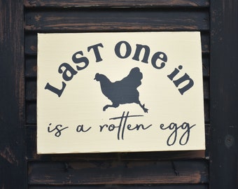 May include: A white wooden sign with black lettering that reads "Last one in is a rotten egg". A black silhouette of a chicken is in the center of the sign.